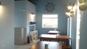 Una habitación con una mesa de madera y bancos, una ventana con vistas, una rueda de barco fijada a la pared y un estante con libros y una pequeña planta.