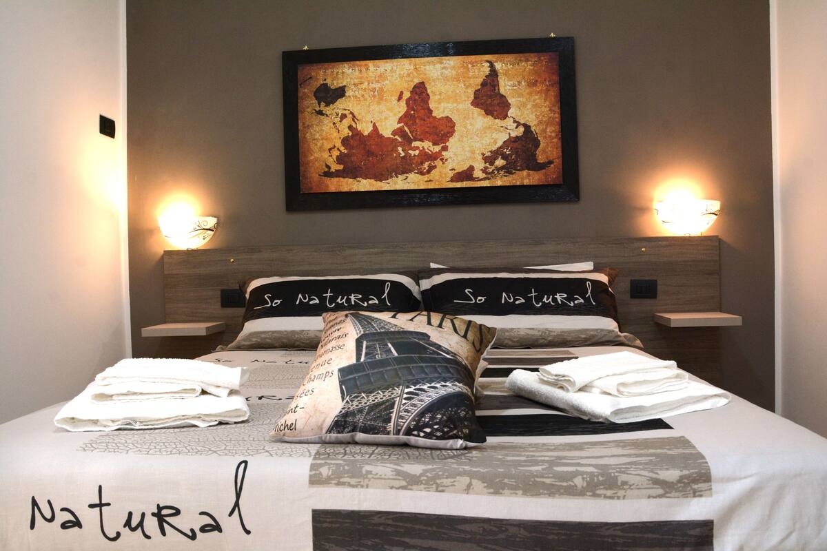 A neatly made bed with pillows labeled "So Natural," a framed map on the wall, and two bedside lamps.
