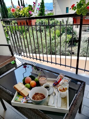 Un balcón con una mesa dispuesta para el desayuno, que incluye pan, frutas, cereales y una taza de café.