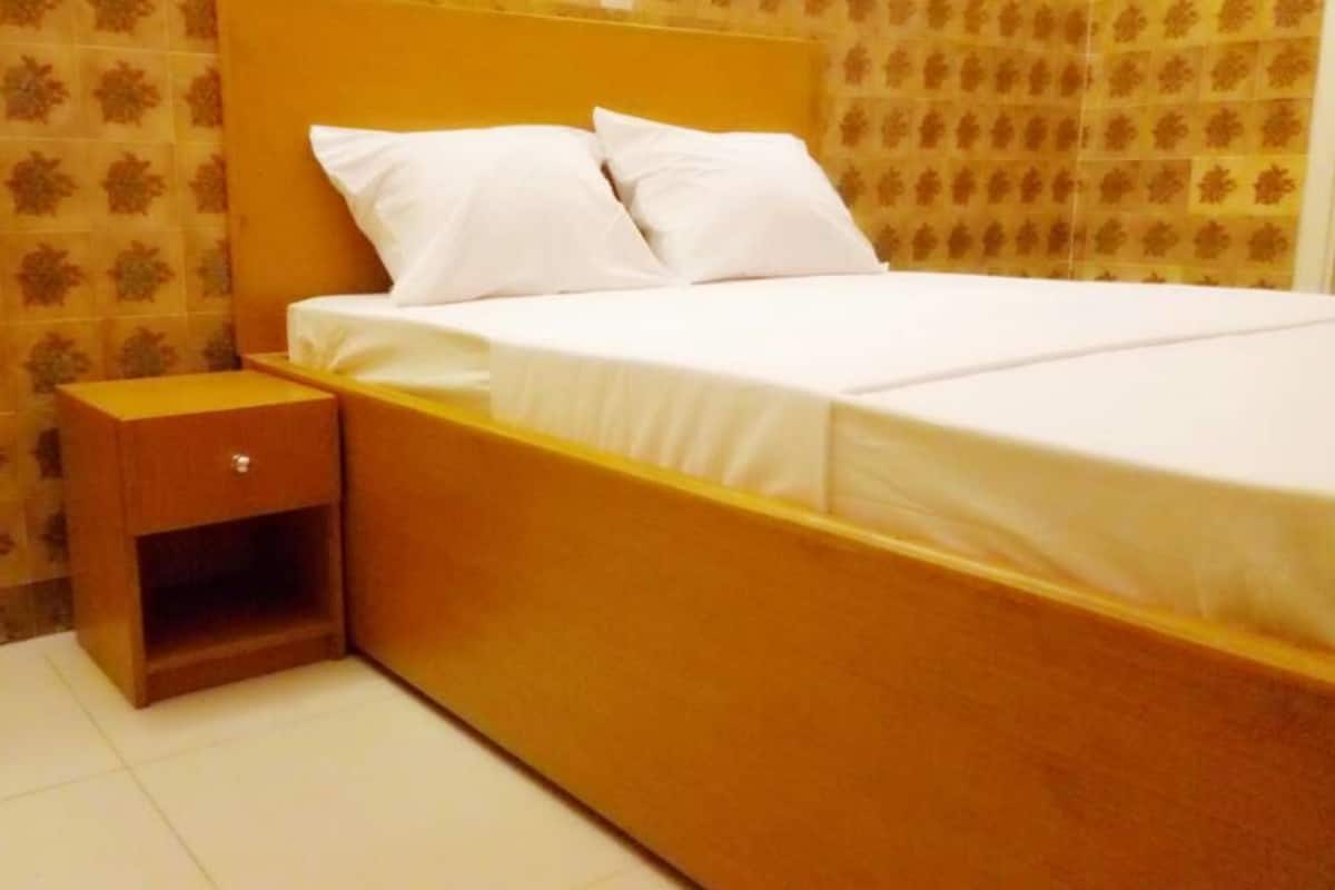 A single bed with white bedding and pillows, a wooden nightstand with a drawer, and a patterned wall.