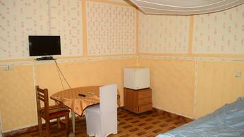 A small, furnished room with a wooden floor, a table with a tablecloth, a chair, a TV on the wall, and a bed.