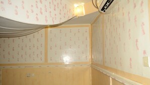 A room with wallpaper featuring a repeated pattern of pink figures, a visible electrical outlet, and a partially visible bed.