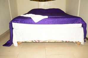 A bed with a purple bedspread and white sheets, a floral-patterned skirt, and a wooden headboard.