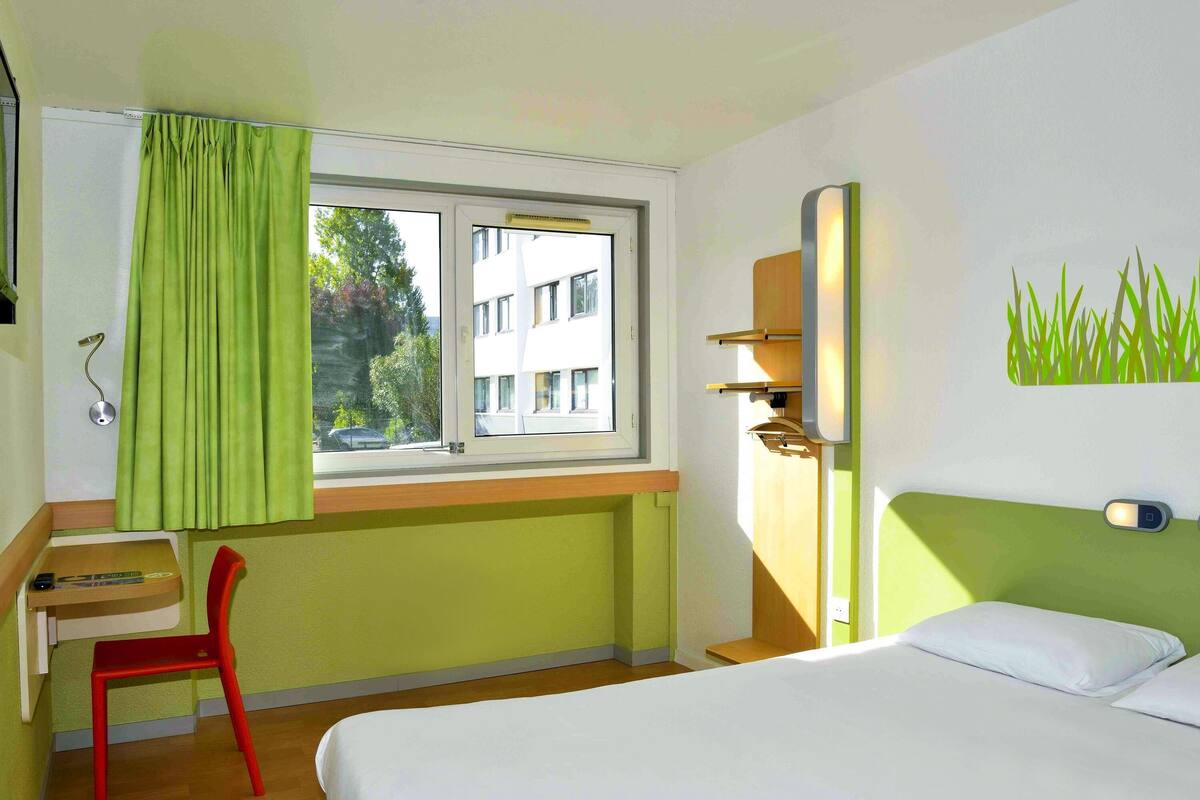 A hotel room with a bed, a desk, a chair, a window with curtains, and a wall-mounted shelf.