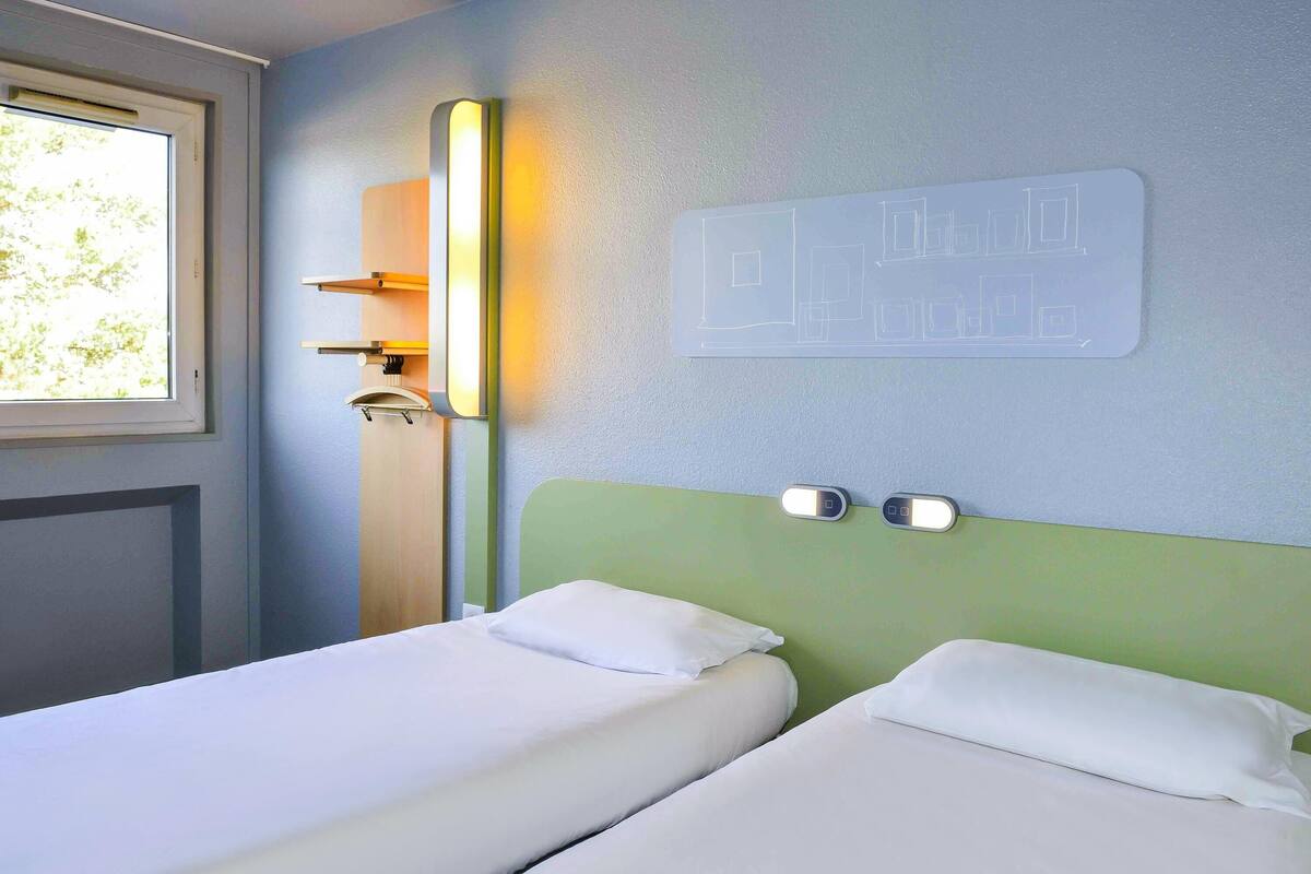 A hotel room with two single beds, a window, a wall-mounted light, and a wall decoration.