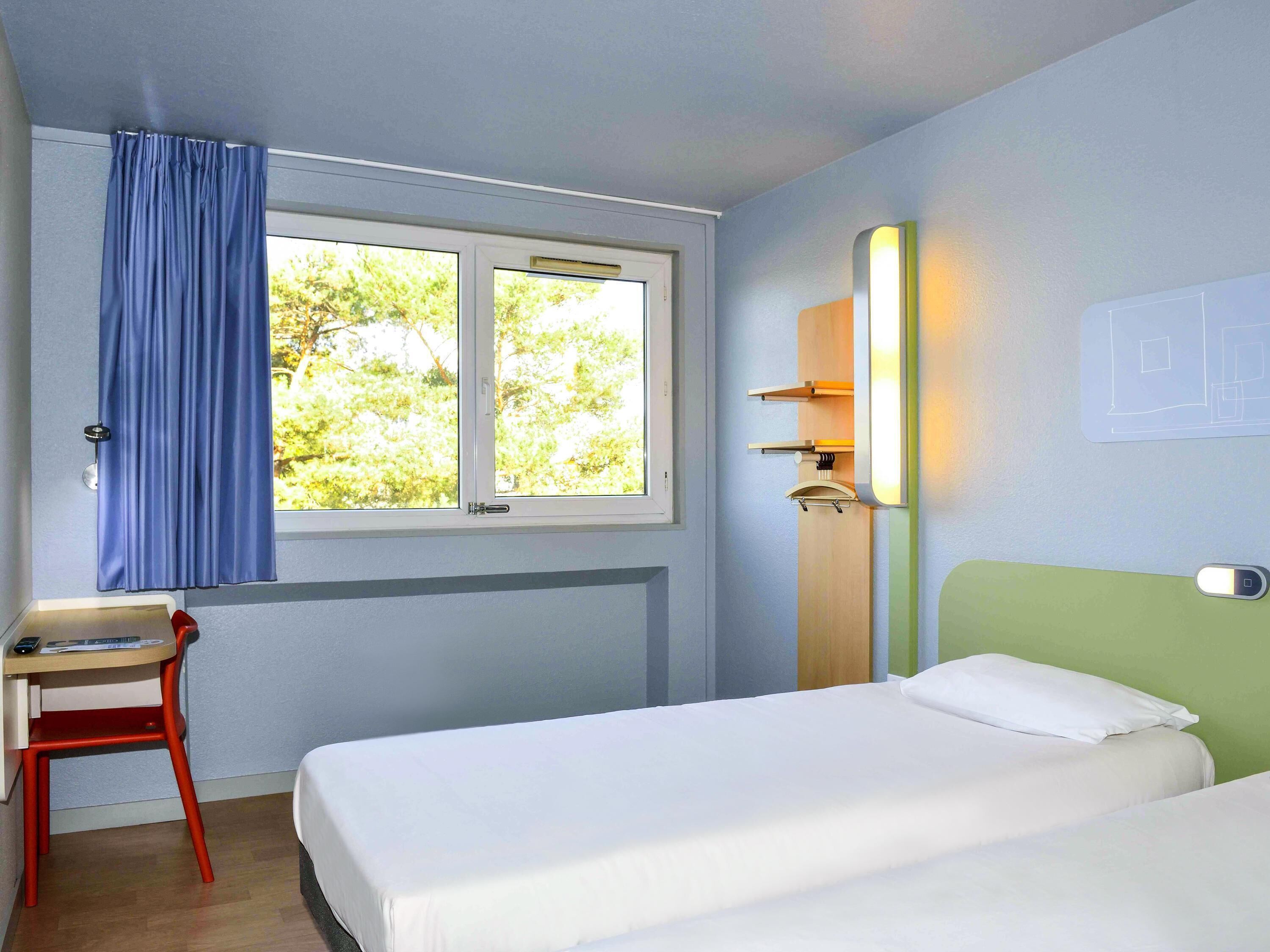 A room with a bed, a desk, a chair, a window with curtains, and a wall-mounted light fixture.