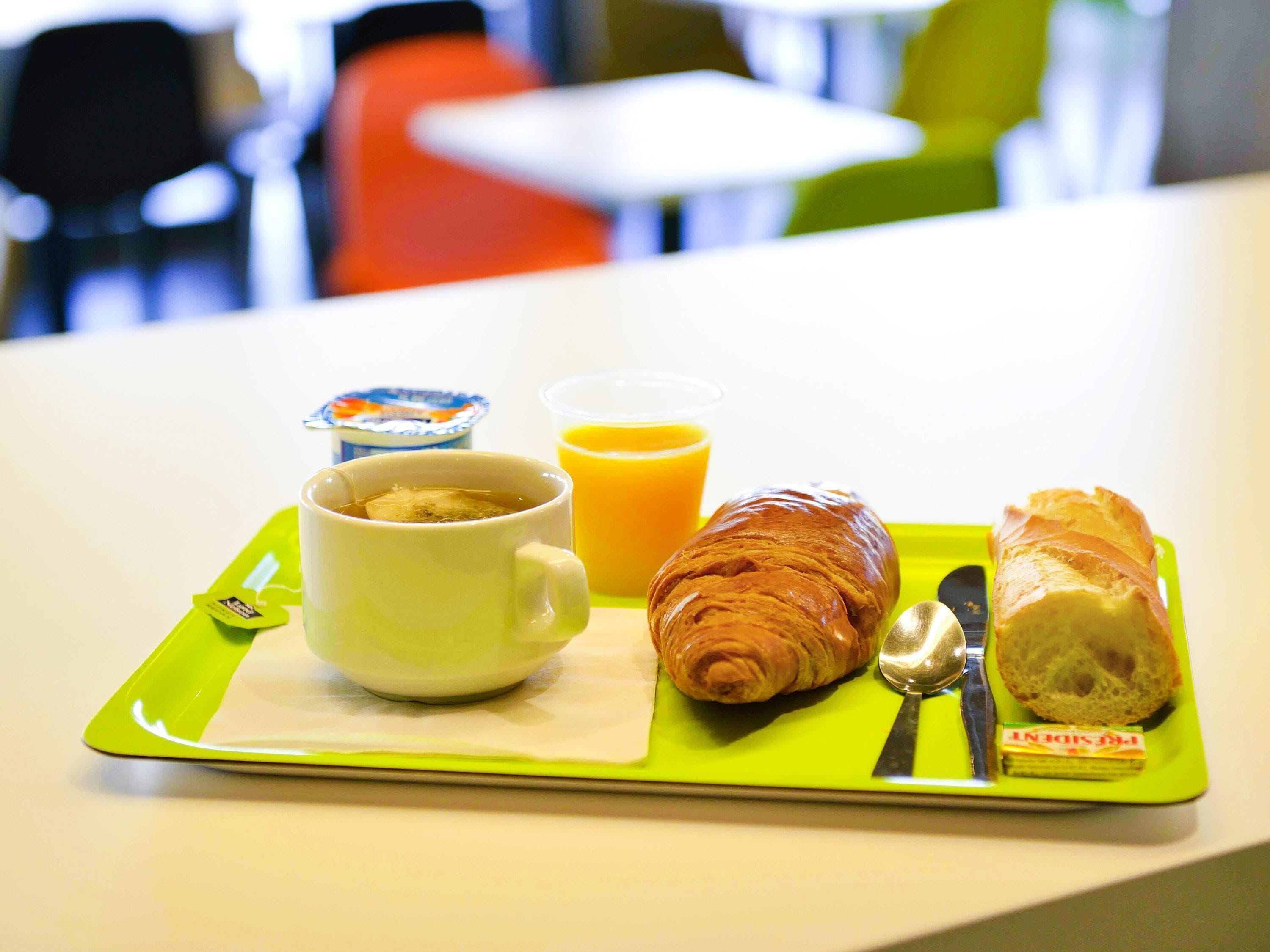 Aamiastarjotin, jossa on kuppi teetä, croissant, viipale leipää, lusikka ja mehupakkaus vihreällä alustalla.