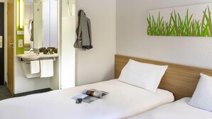 Una habitación de hotel con cama, una mesita de noche con una revista, un espejo, un perchero para toallas y una obra de arte en la pared.