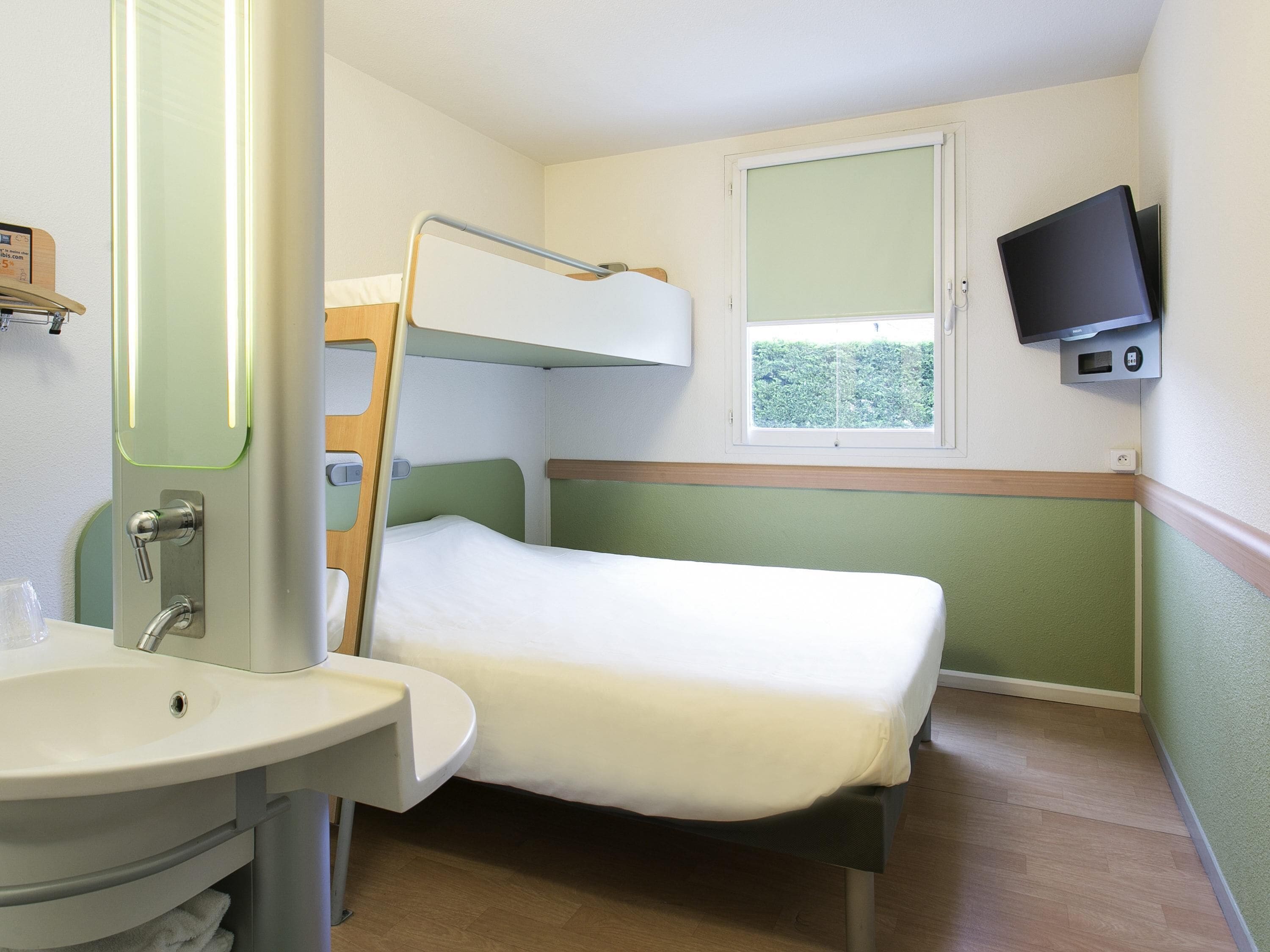 Ein kleines Zimmer mit einem Etagenbett, einem Nachttisch, einem Spiegel, einem Waschbecken und einem an der Wand befestigten Fernseher.