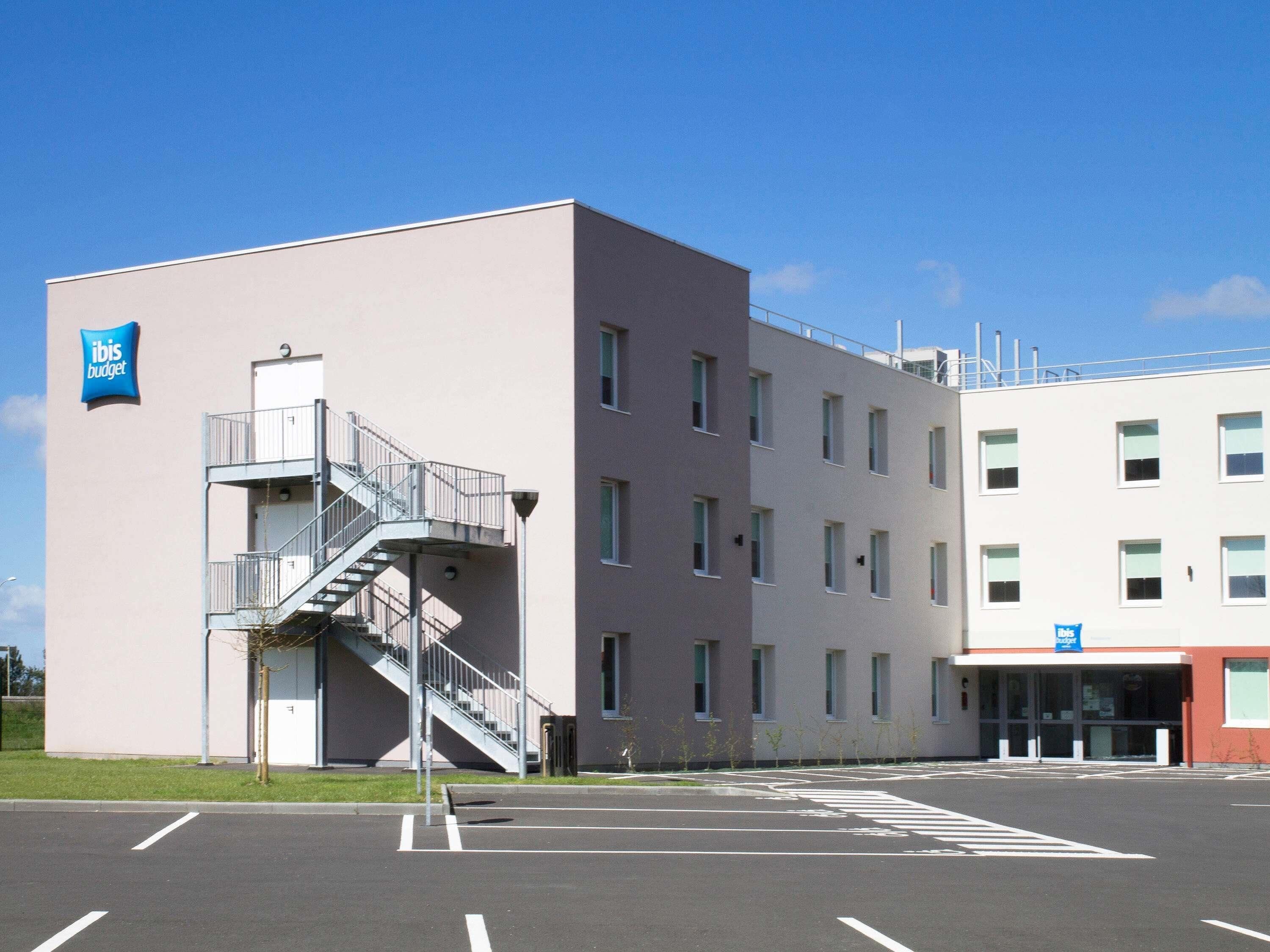 Un edificio moderno con fachada plana, una escalera que conduce a un nivel superior y un estacionamiento con espacios delimitados.
