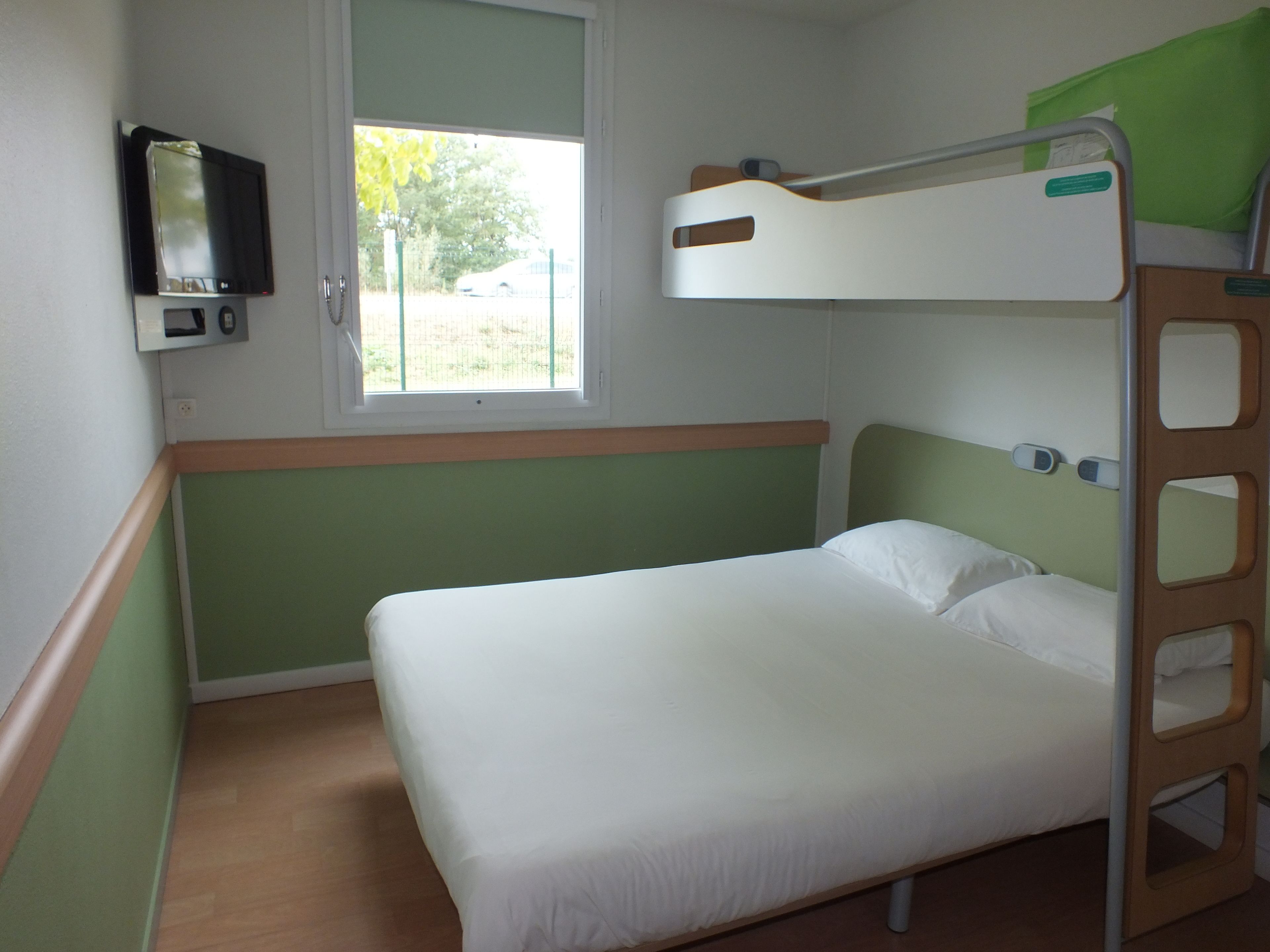 A bunk bed with a green headboard, a bedside table, a TV mounted on the wall, and a window with a view of the outdoors.