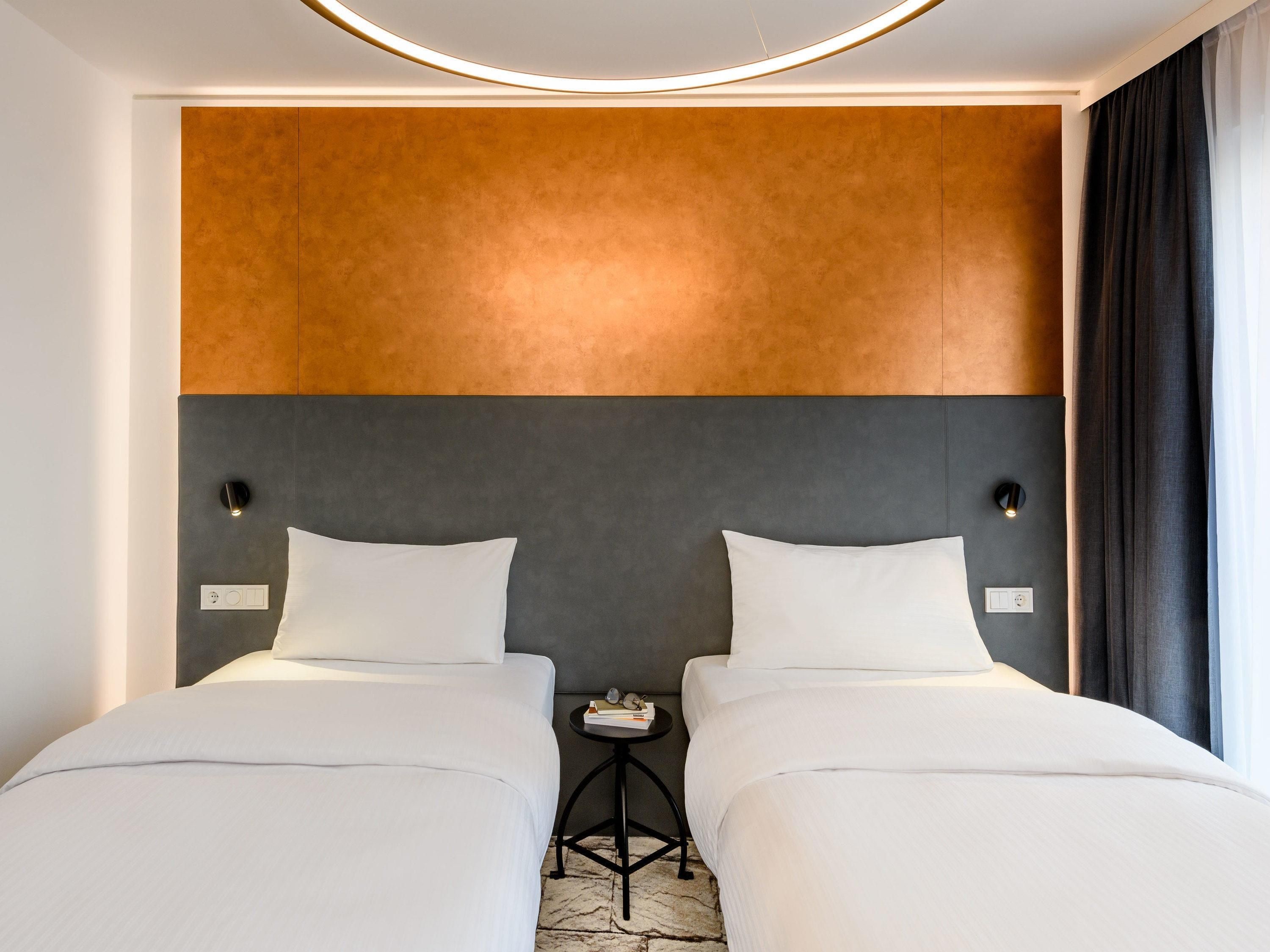 A hotel room with a modern design, featuring two beds with white linens, a grey and brown headboard, and a small bedside table with a lamp.