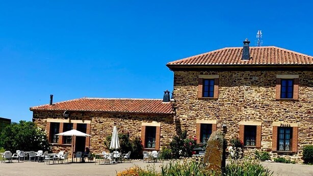 Un bâtiment en pierre avec un toit recouvert de tuiles, un espace extérieur aménagé pour s’asseoir et un ciel d’un bleu limpide.