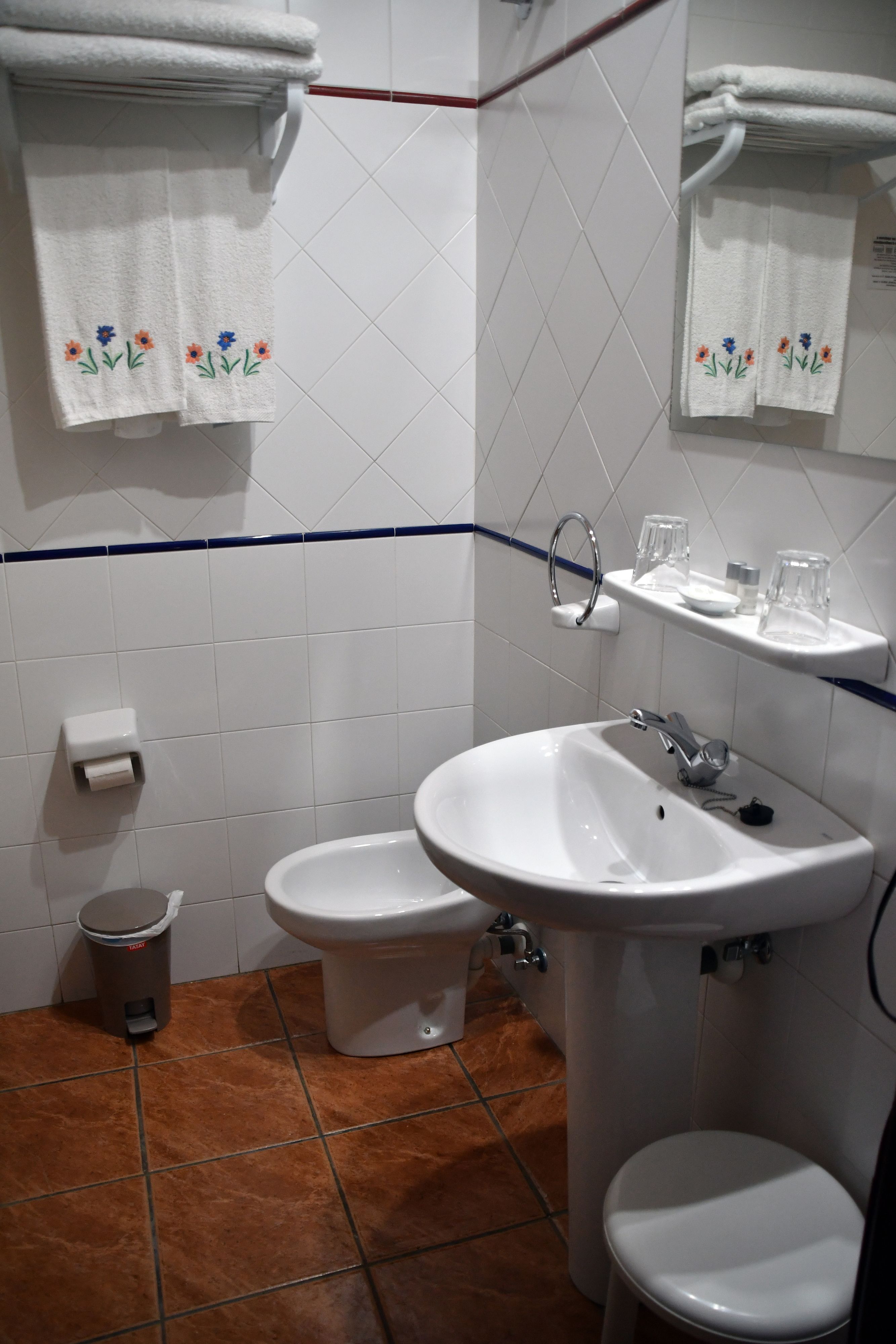 Un baño con un lavamanos blanco, dos inodoros, un cubo de basura y toallas con diseños florales.