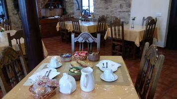 Uma área de refeições com uma mesa posta para dois, apresentando uma parede de pedra ao fundo, cadeiras de madeira e diversos utensílios de mesa e cestas de pão.