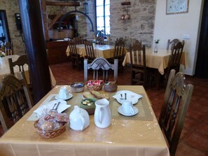 A dining area with a table set for two, featuring a stone wall backdrop, wooden chairs, and a variety of tableware and bread baskets.
