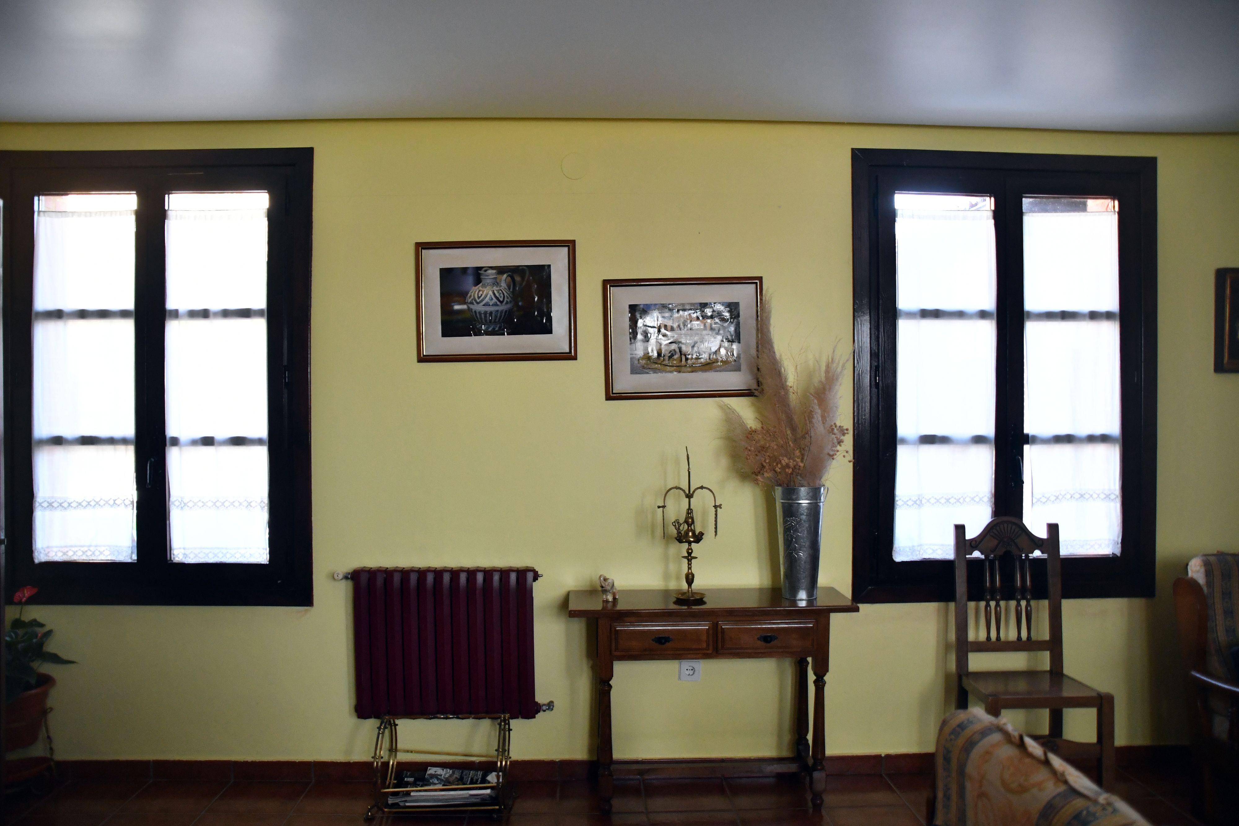 Una habitación con dos ventanas, un radiador, una silla de madera, una mesita y dos cuadros en la pared.