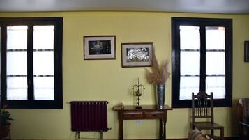 Una habitación con dos ventanas, un radiador, una silla de madera, una mesita y dos cuadros en la pared.