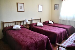 A hotel room with three beds, each with a wooden headboard and purple bedding. There are framed pictures on the walls and a window with curtains.