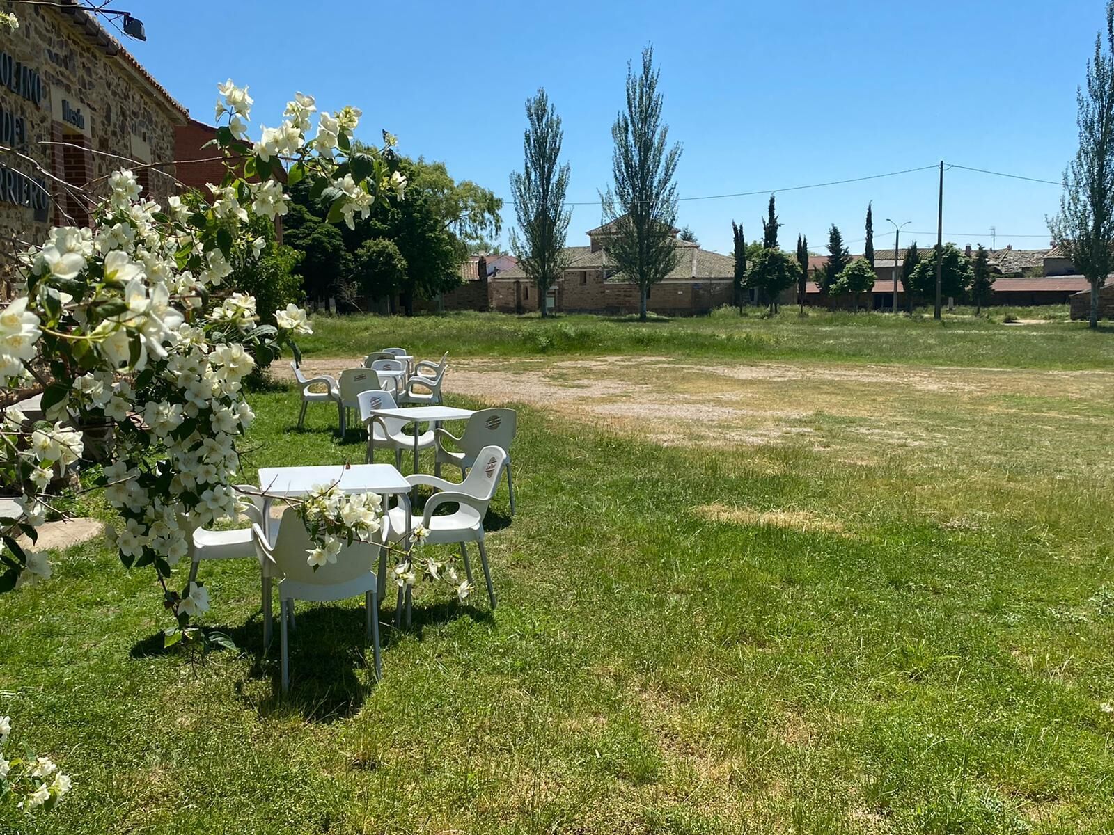 An outdoor seating area with white tables and chairs, surrounded by grass and trees.