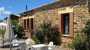 Un espace extérieur aménagé avec des chaises et des tables en plastique blanc, un bâtiment en pierre avec une partie en briques, et une enseigne sur laquelle on peut lire « San Miguel ».