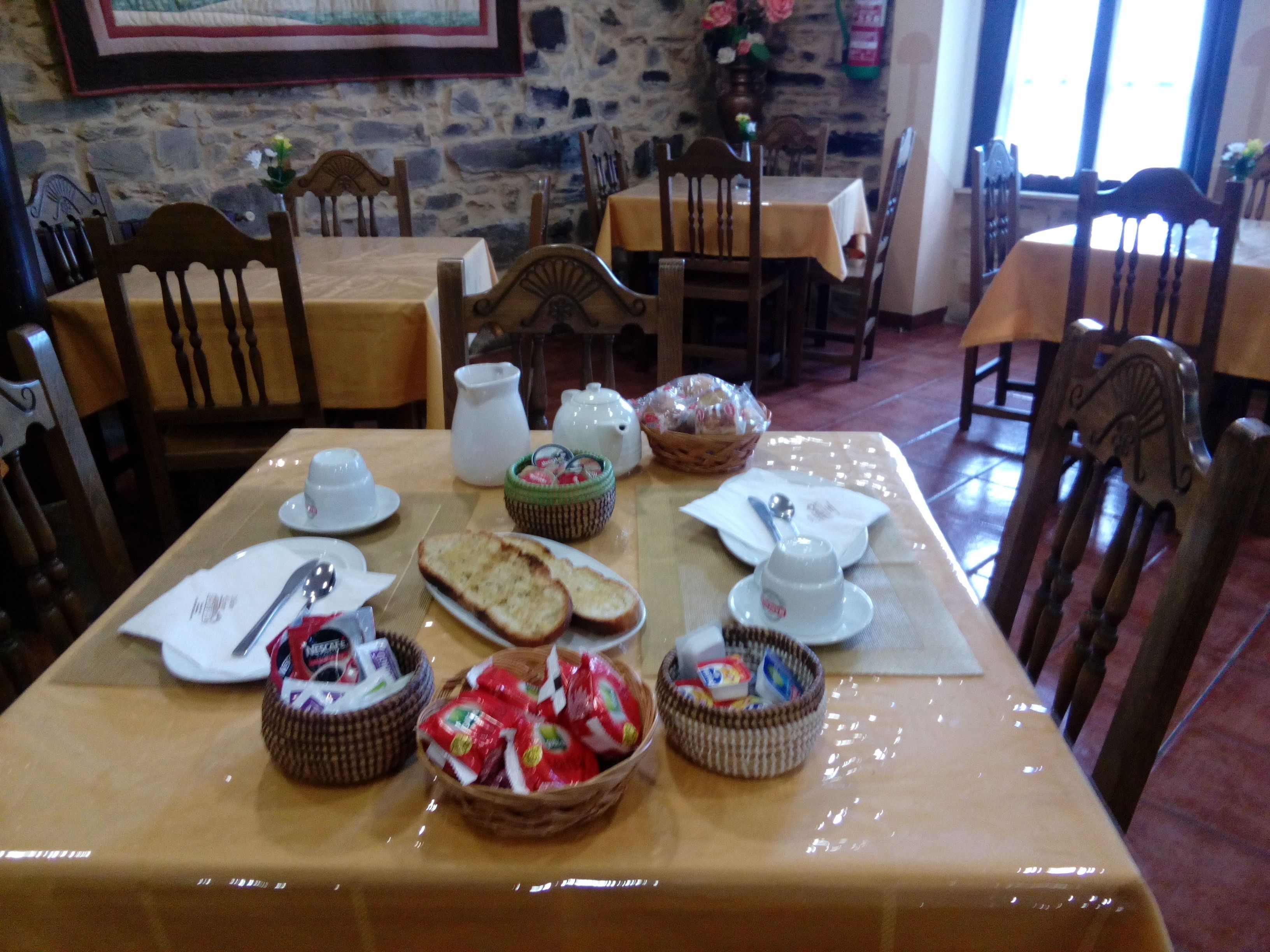 A dining area with a stone wall, wooden chairs, a table set with bread, tea cups, and baskets of snacks.