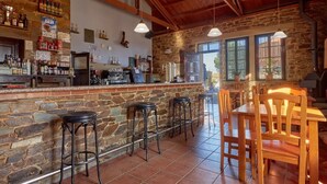 Un coin bar au style rustique, avec des murs en pierre, des poutres en bois au plafond et un sol carrelé. On y trouve des tabourets de bar et une table en bois avec des chaises.