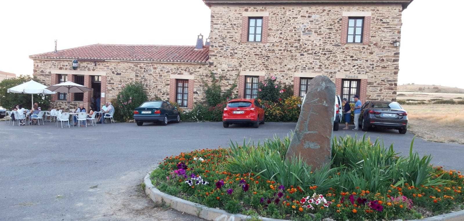 Un bâtiment en pierre avec un toit en tuiles, un espace de stationnement et un parterre fleuri où poussent différentes espèces de fleurs.