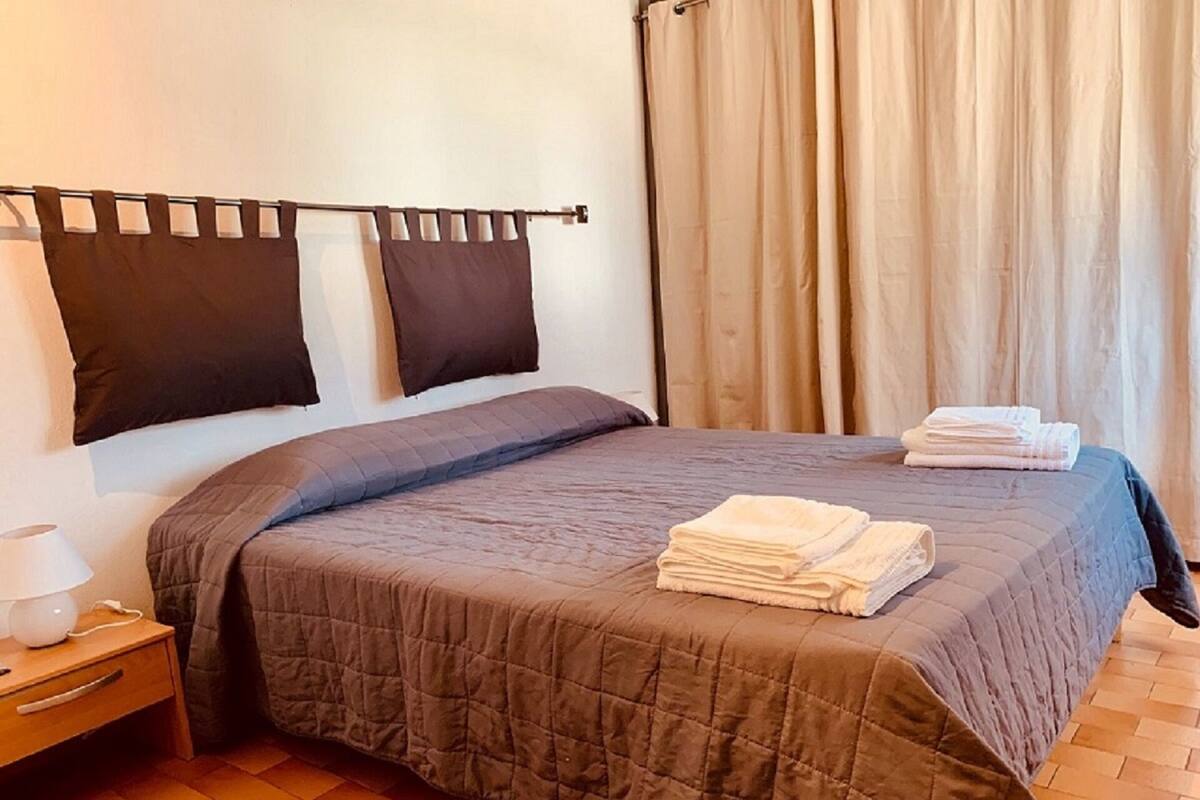 Un lit bien fait, recouvert d’une couverture sombre, une lampe de chevet et une pile de serviettes pliées sur la table de chevet.