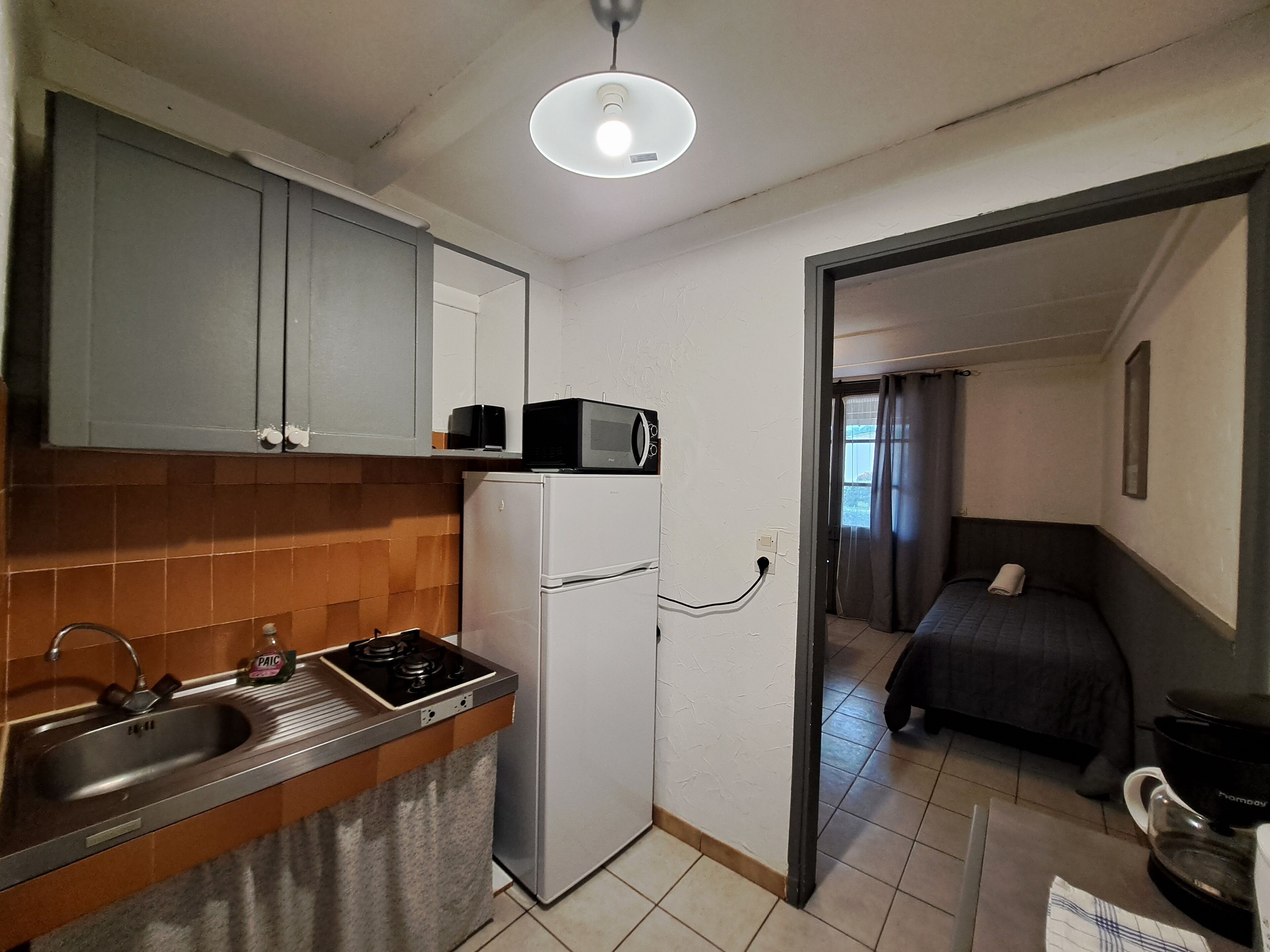 A compact kitchen with a sink, stove, and microwave. A bed and a coffee maker are visible in the adjacent room.