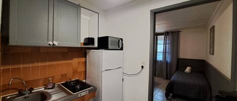 A compact kitchen with a sink, stove, and microwave. A bed and a coffee maker are visible in the adjacent room.