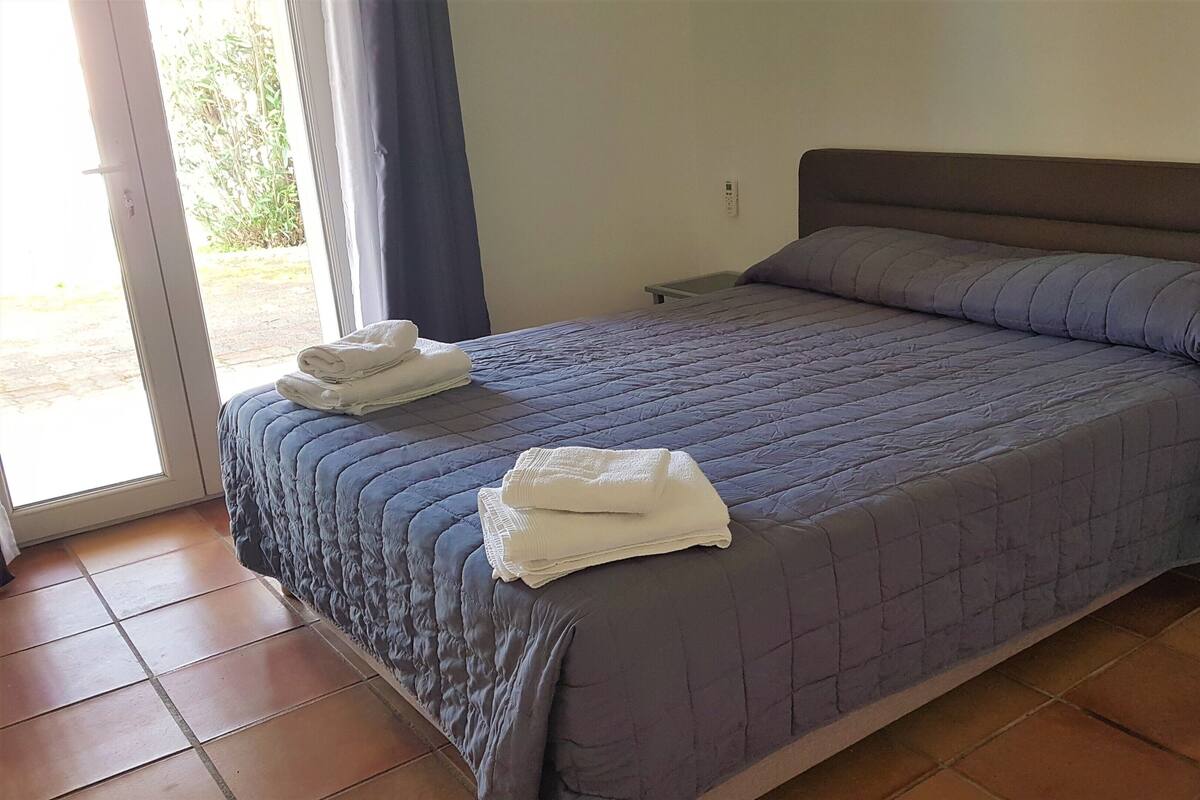 Un lit bien fait, recouvert d’une courtepointe bleue, avec deux serviettes blanches et une petite table de chevet surmontée d’une lampe.