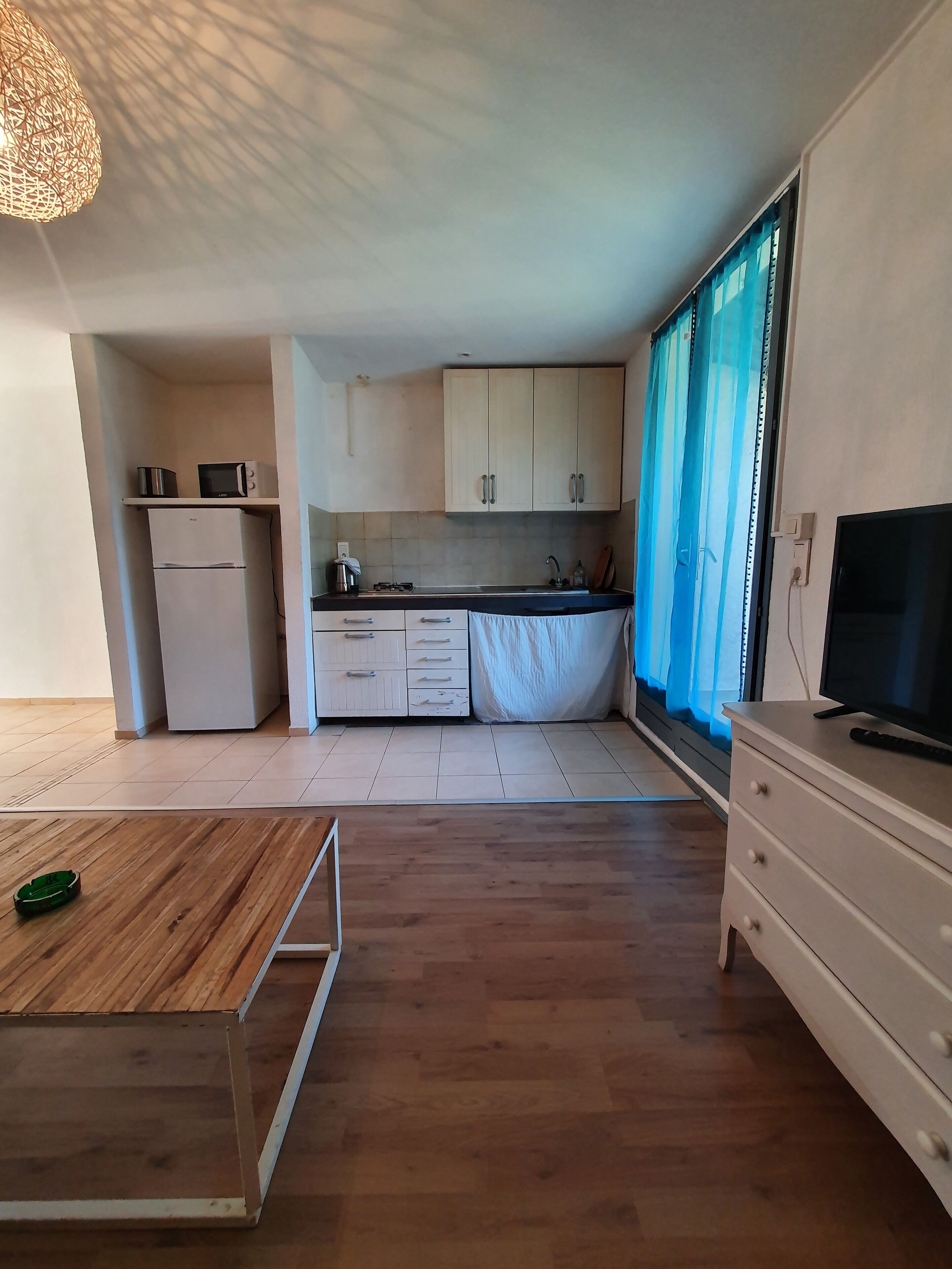 A compact kitchen with a refrigerator, microwave, and stove, a wooden dining table, and a TV on a dresser.