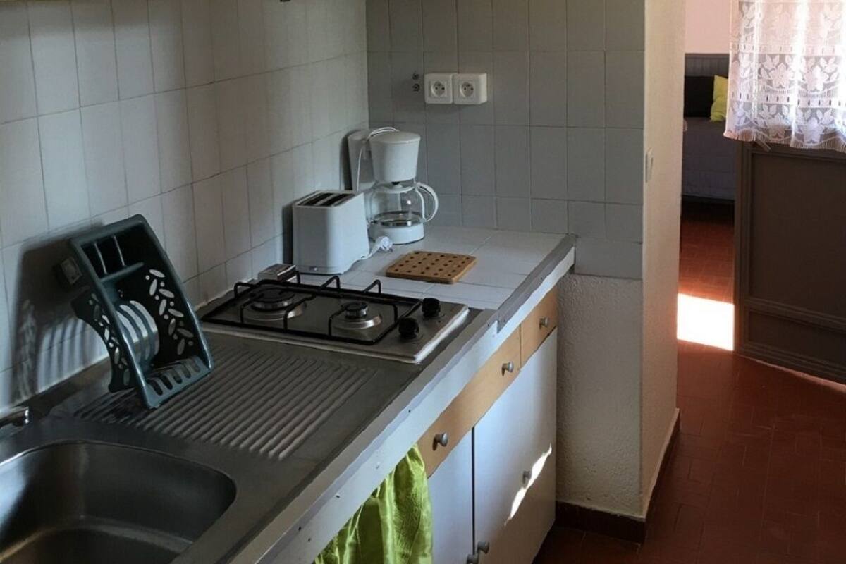 Une cuisine compacte équipée d’une cuisinière à gaz, d’un évier et d’un four à micro-ondes. Une fenêtre avec un rideau laisse entrer la lumière, et la cuisine est dotée d’un dosseret carrelé.