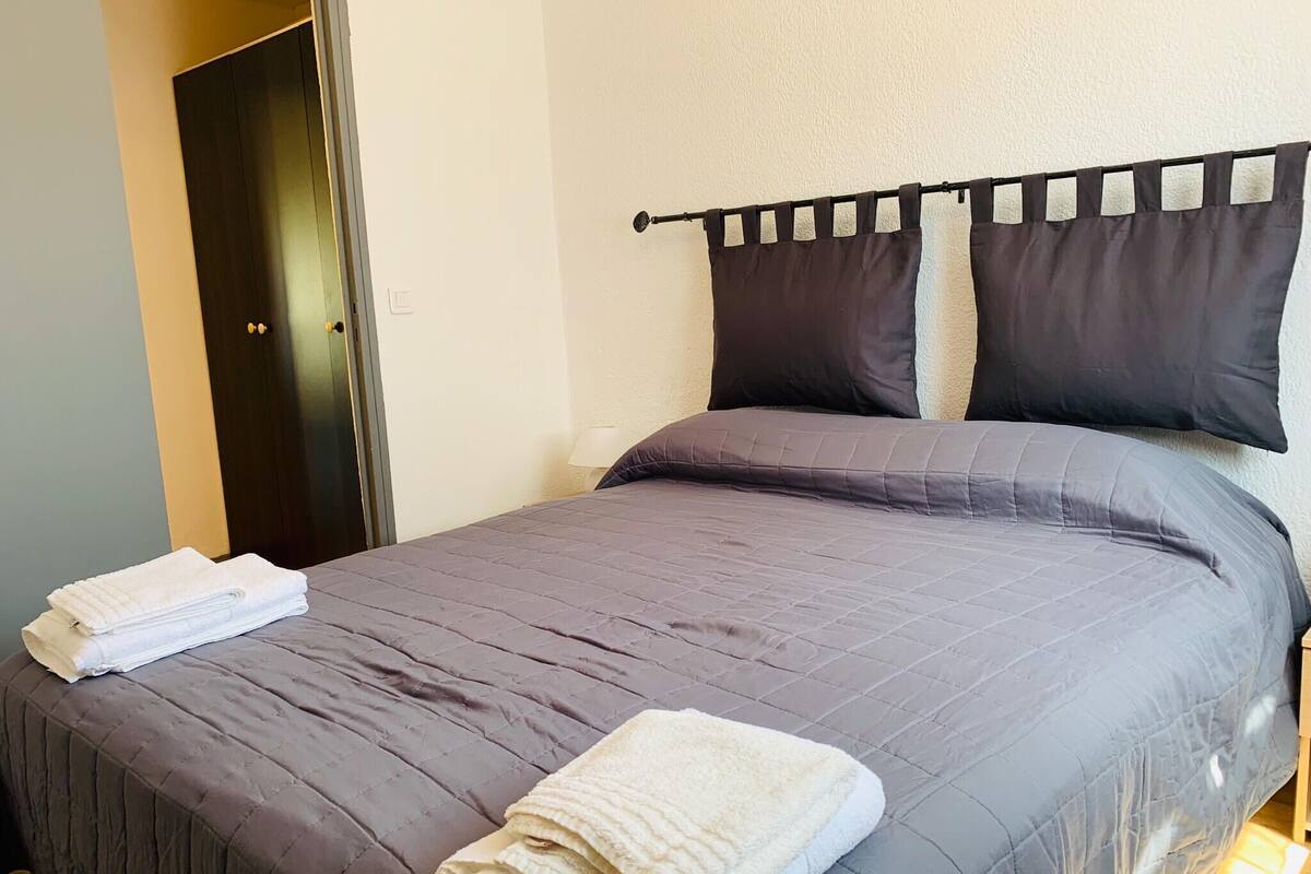 Un lit bien fait, avec une couette et des oreillers gris, une pile de serviettes blanches posée sur le pied de lit, et une porte sombre sur le côté gauche.