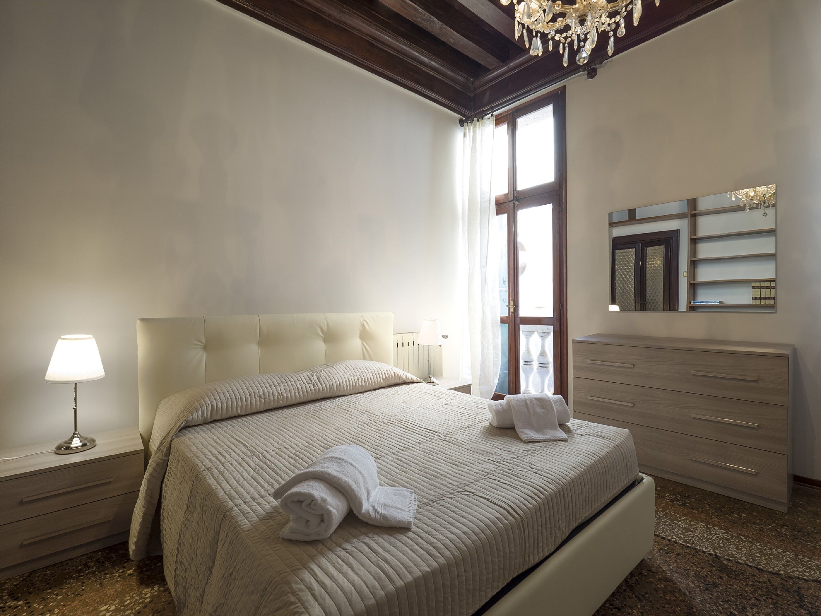 A bedroom with a large bed, a nightstand with a lamp, a dresser, and a mirror on the wall.