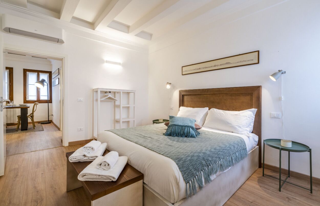 Una camera da letto moderna con una testiera in legno, un letto con lenzuola bianche e azzurre, un comodino con asciugamani e una piccola abat-jour.
