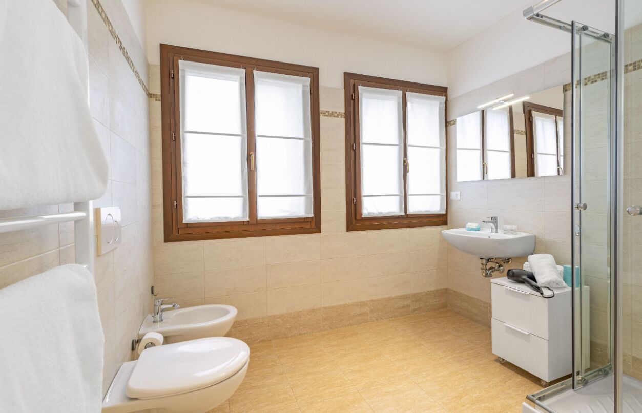 Een badkamer met twee toiletten, een wastafel, een douchecabine van glas en twee ramen met houten kozijnen.
