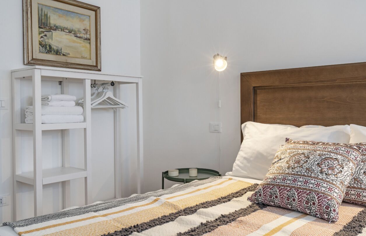 A neatly arranged bedroom with a bed, a side table, a hanging wardrobe, and a painting on the wall.