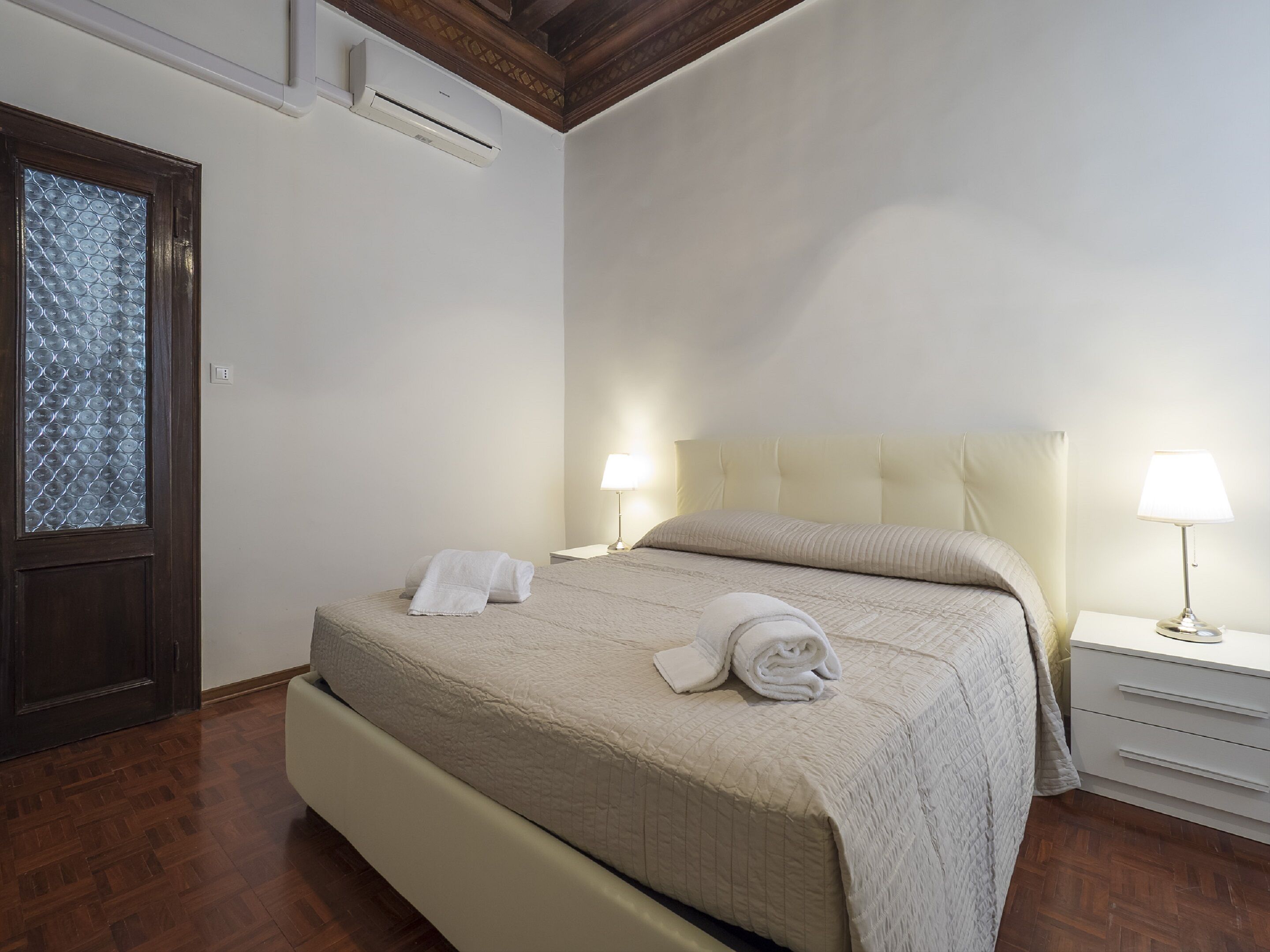 A hotel room with a bed, two bedside tables with lamps, a wooden door with a frosted glass panel, and an air conditioning unit on the wall.