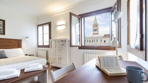 A hotel room with a bed, a table with a book and a mug, a window with a view of a building, and a radiator. - San Marco Schiavoni Apartments (Venice)