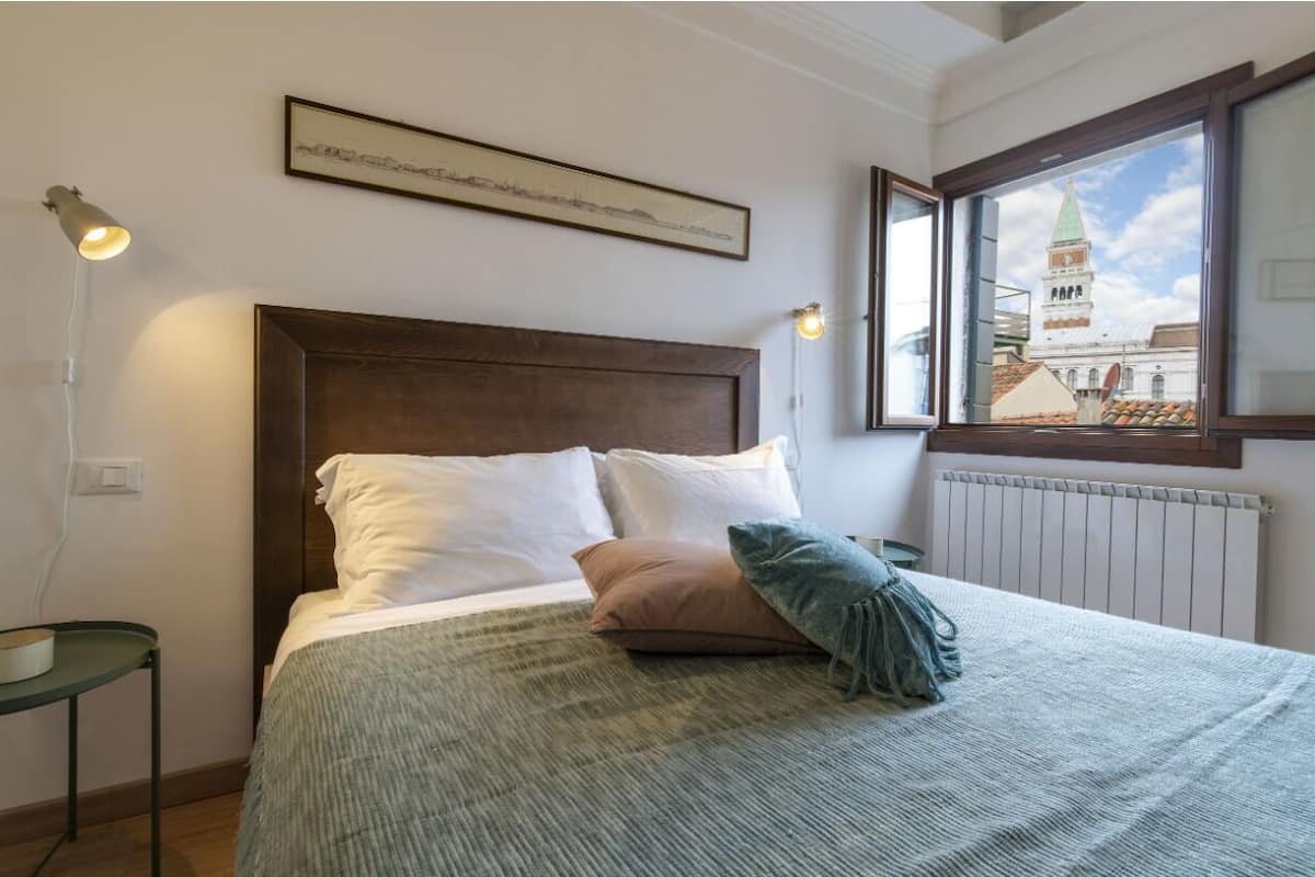 A neatly made bed with a headboard, a framed picture above it, a bedside lamp, and a window with a view of a building.