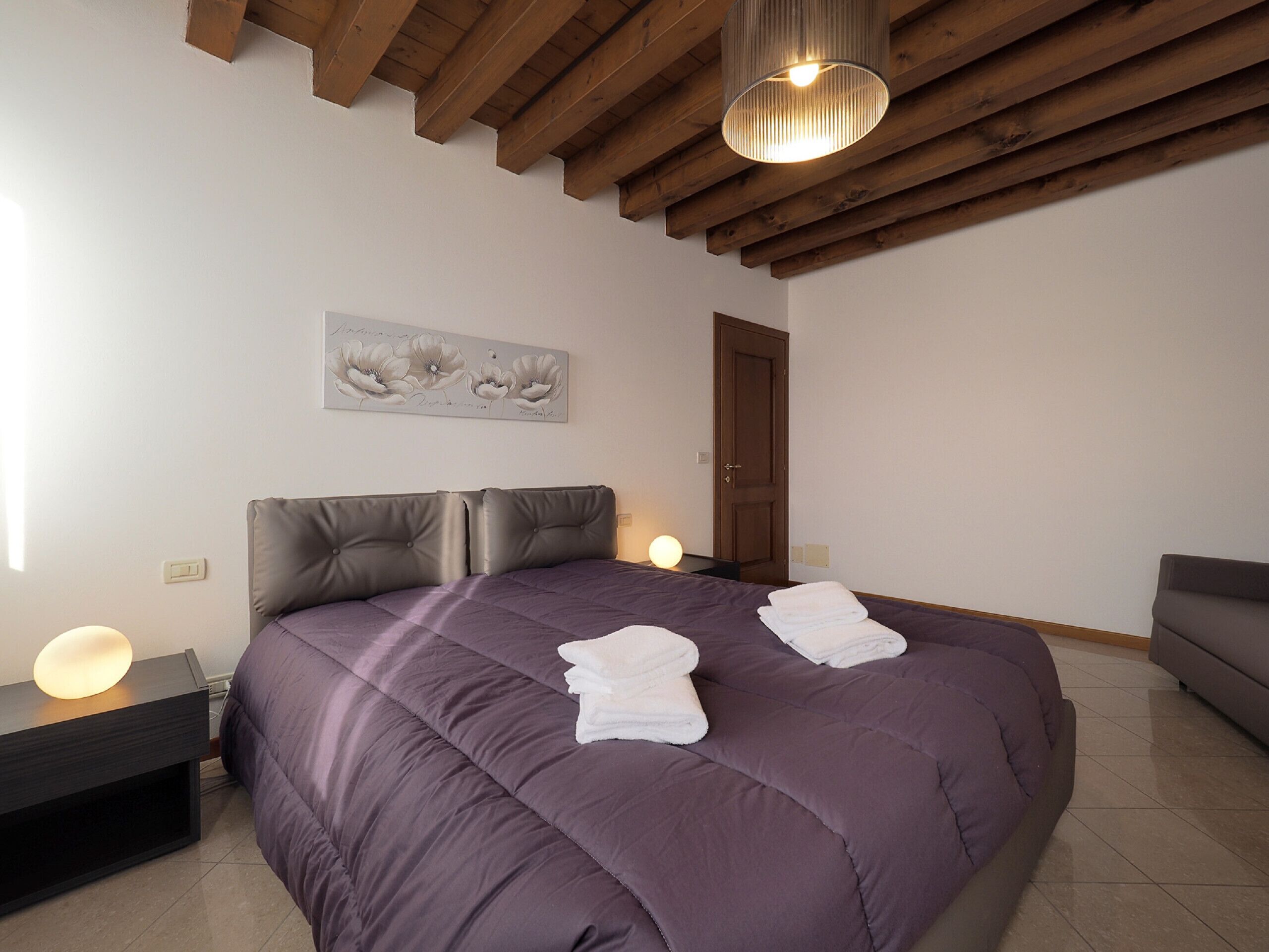 A bedroom with a large bed, a nightstand, a bedside table with a lamp, a door, and a wall-mounted artwork.