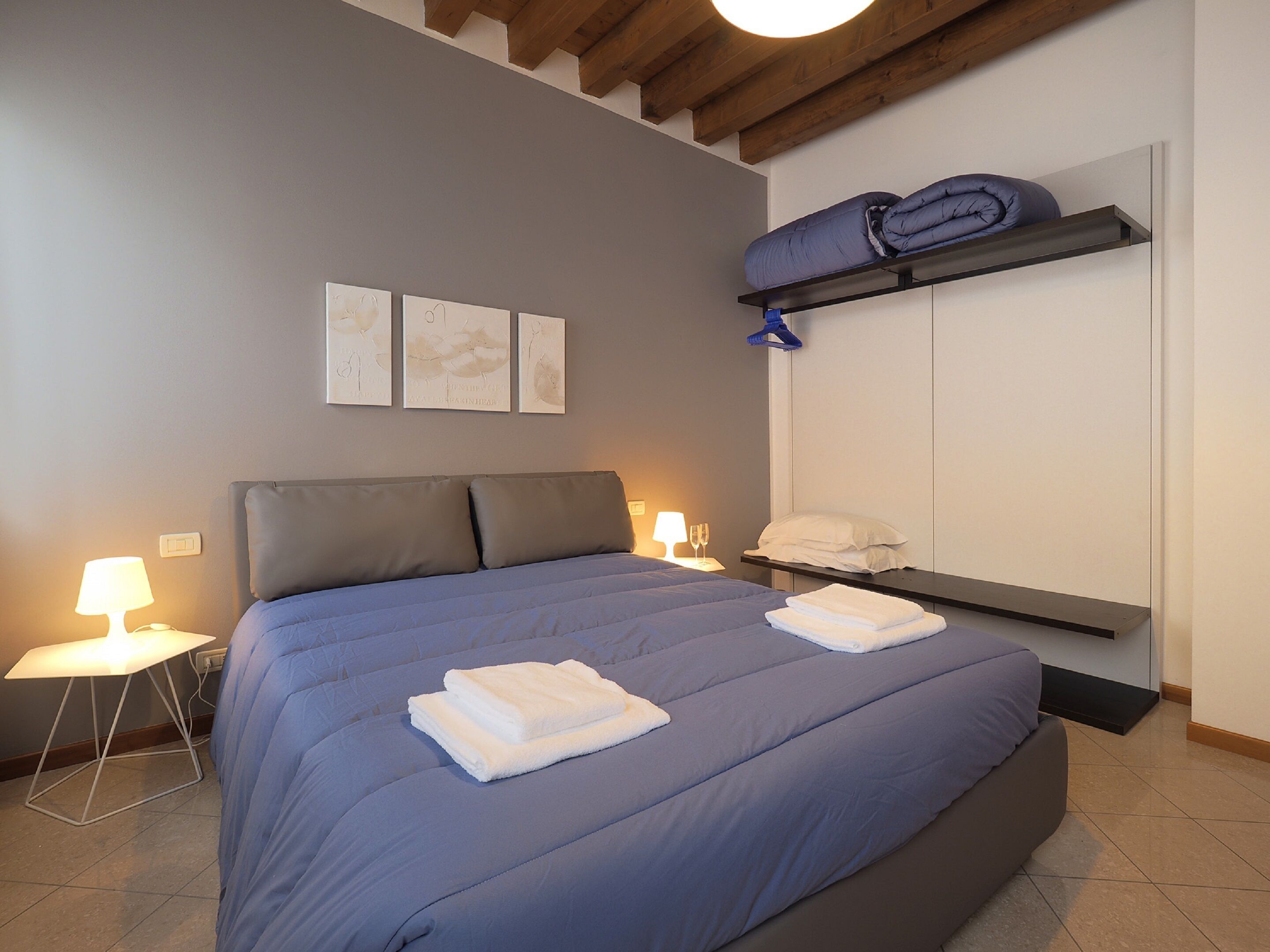 A neatly made bed with blue bedding and white towels, a bedside table with a lamp, and a wall-mounted shelf with folded clothes.