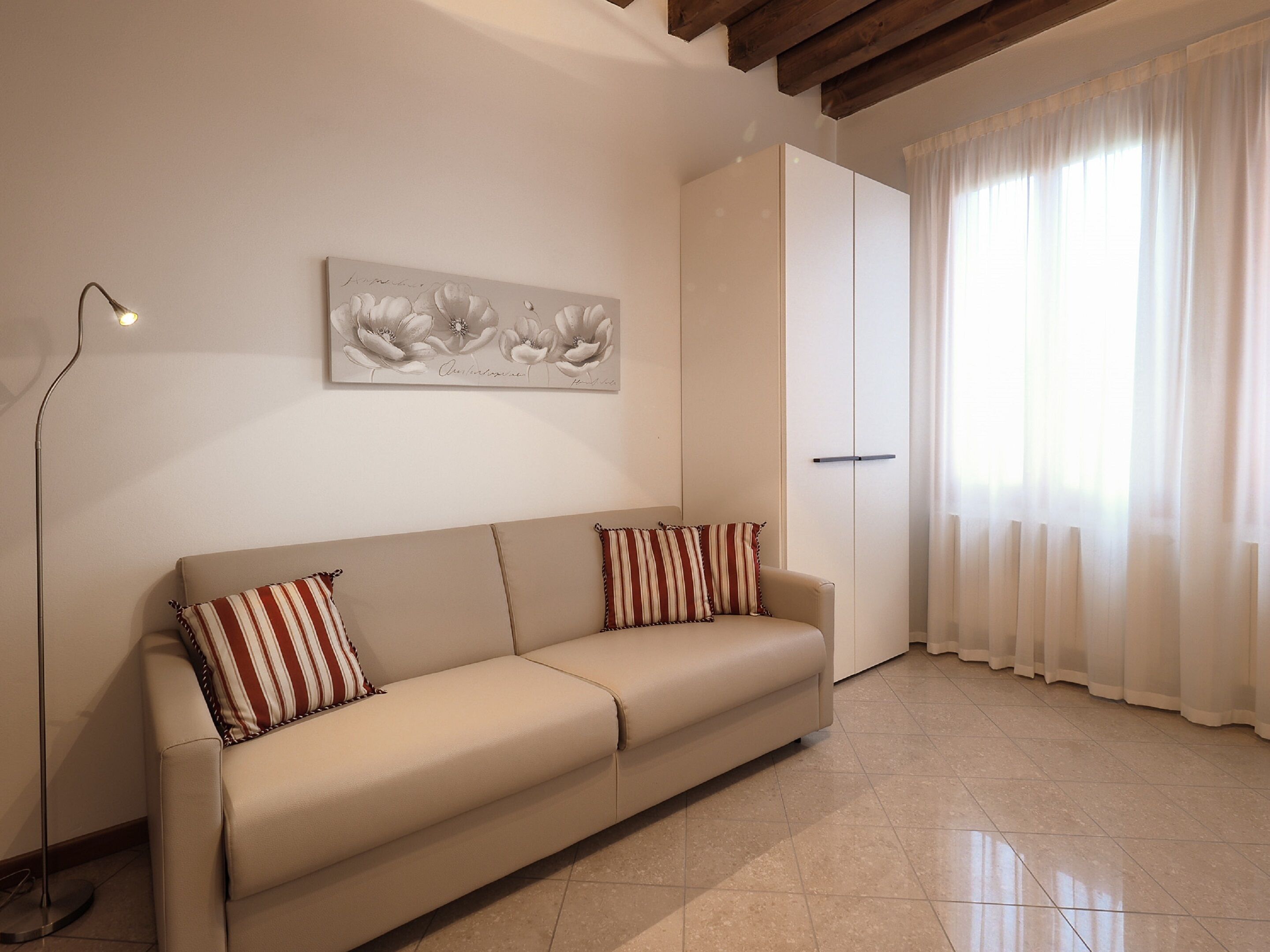 A neutral-toned living room with a beige sofa, striped pillows, a floor lamp, a large window with sheer curtains, and a wall-mounted artwork.