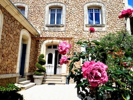 Un edificio de piedra con una entrada central, escalones que llevan a ella y un pequeño jardín con flores rosas.