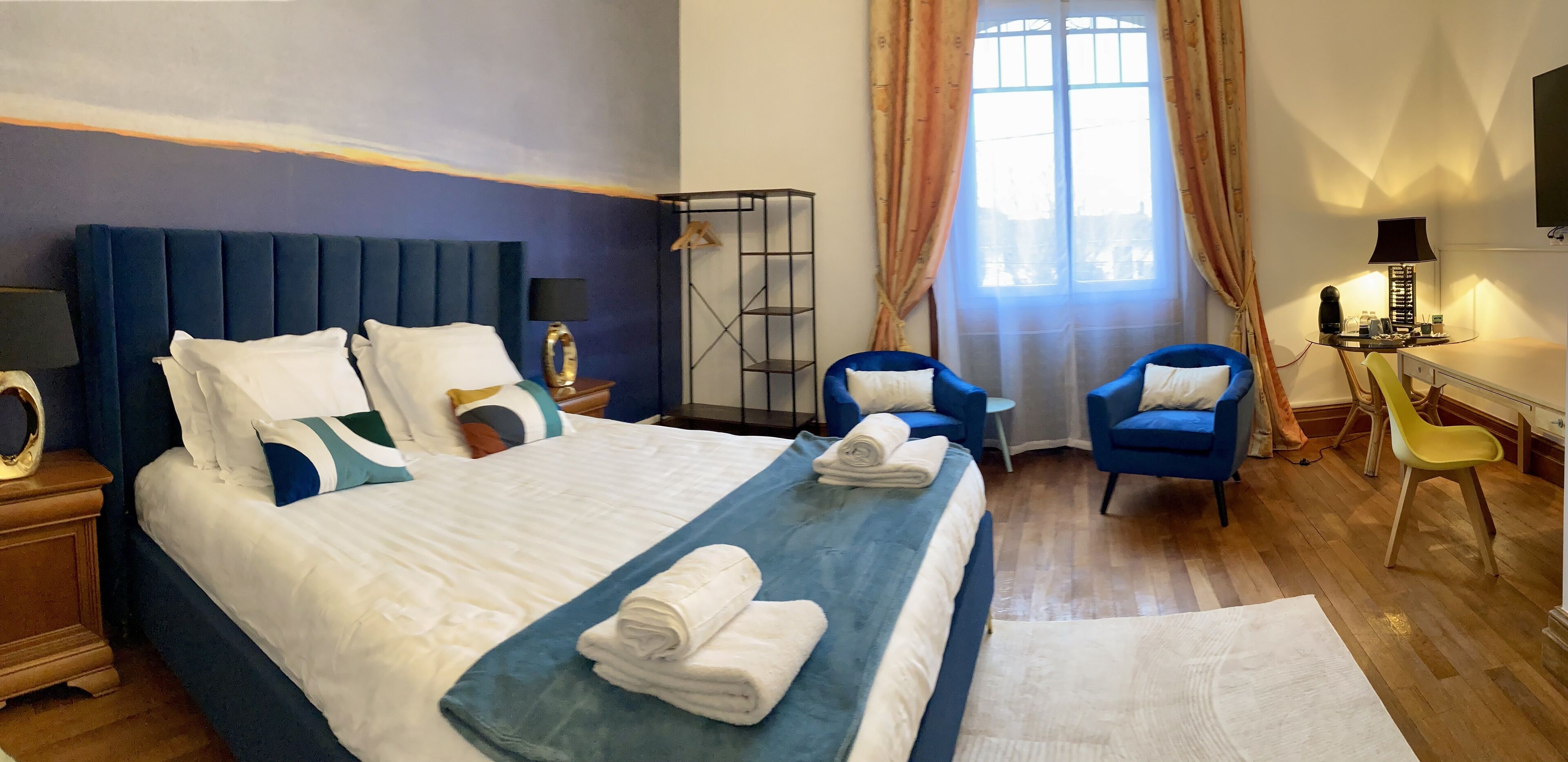 A hotel room with a large bed, two blue armchairs, a desk with a lamp, and a TV on the wall.
