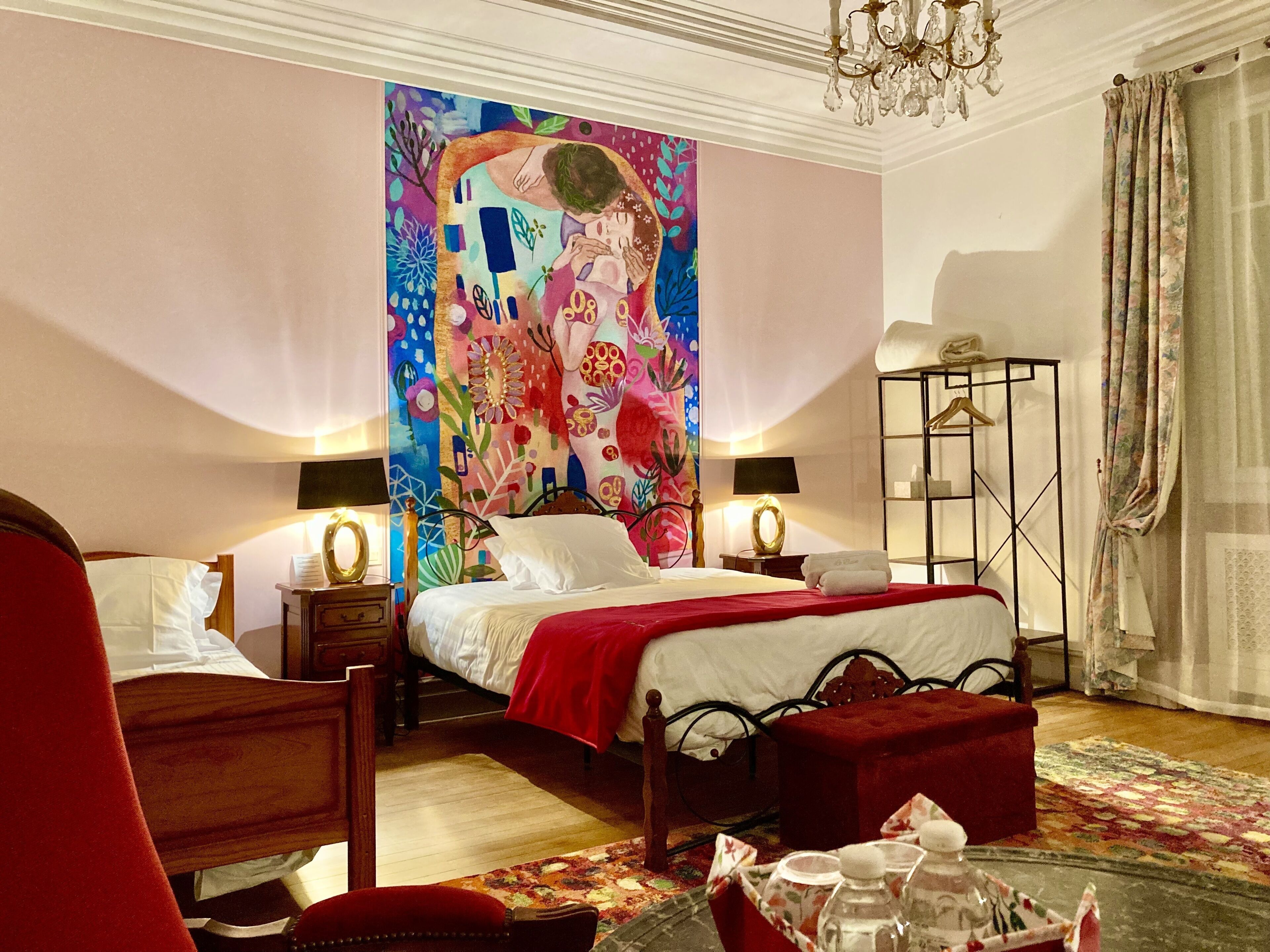A hotel room with a large colorful painting on the wall, a bed with a red blanket, a bedside table with a lamp, a chair, and a floor lamp.