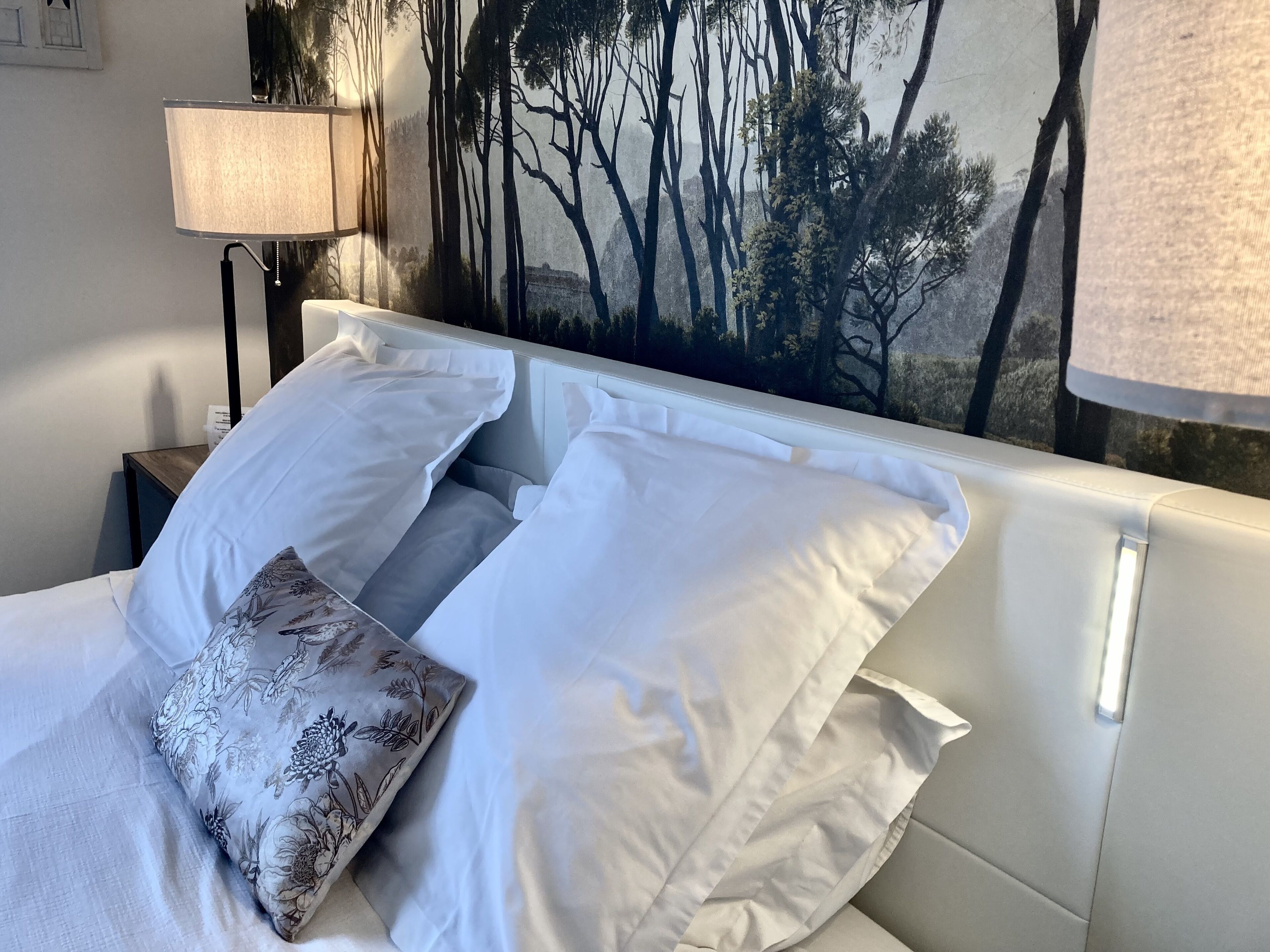 Una cama bien hecha con sábanas blancas y un cojín floral, una lámpara de pared y un mural con motivos de bosque.