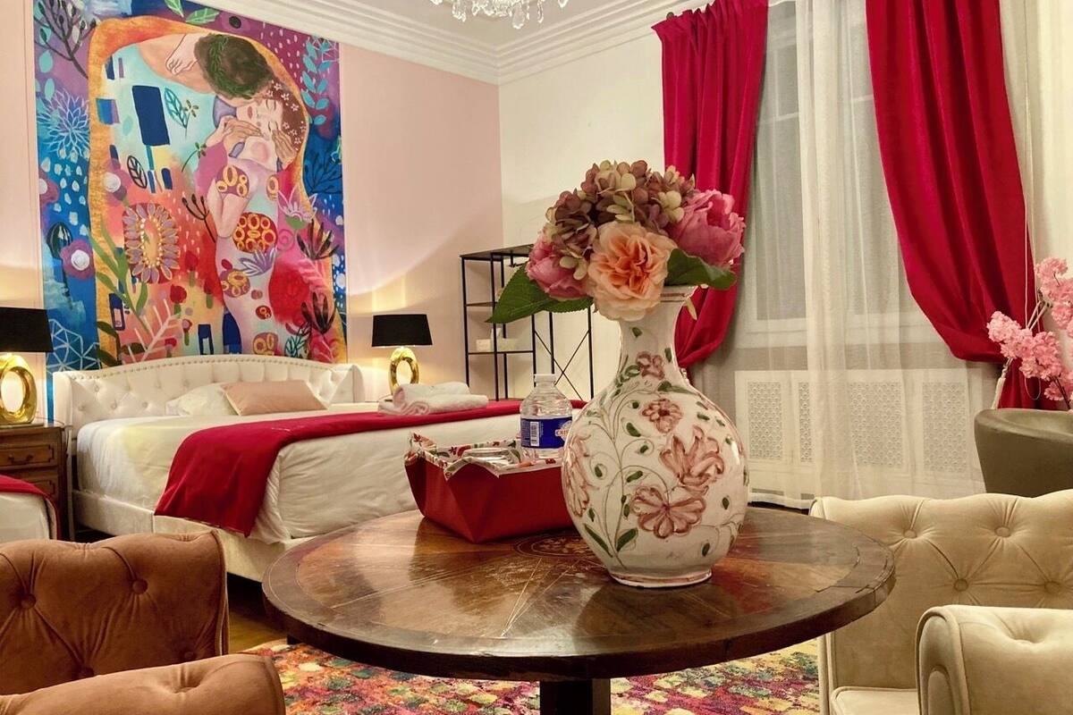 A hotel room with a bed, a round table with a vase, a colorful rug, and a painting on the wall.